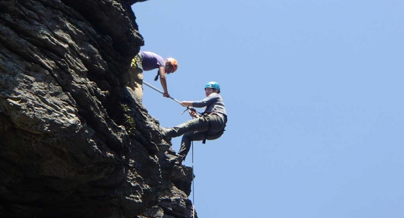 Two people wearing safety gear are secured by ropes near the edge of a cliff. One person appears to be an instructor, giving direction to the other person.